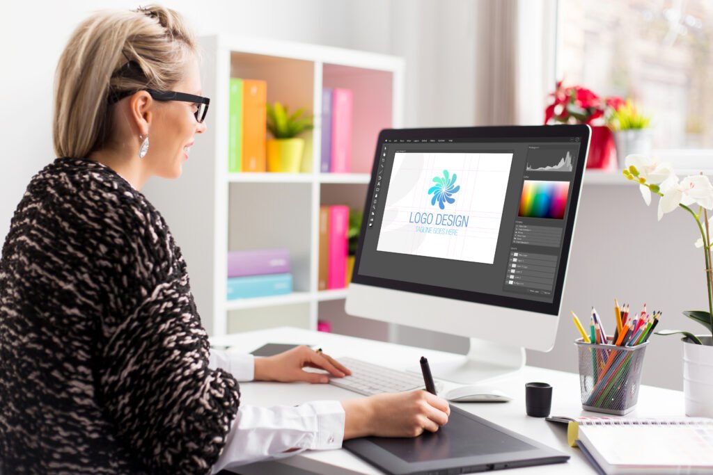 Person sitting at a desk using a digital drawing tablet and computer to design a logo, in a bright home office with shelves, plants, and colorful stationery.