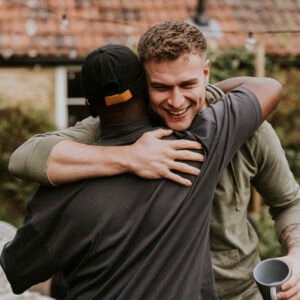 Two friends warmly hugging outdoors in a backyard setting. Natural relaxed environment with greenery and a cozy home in the background.