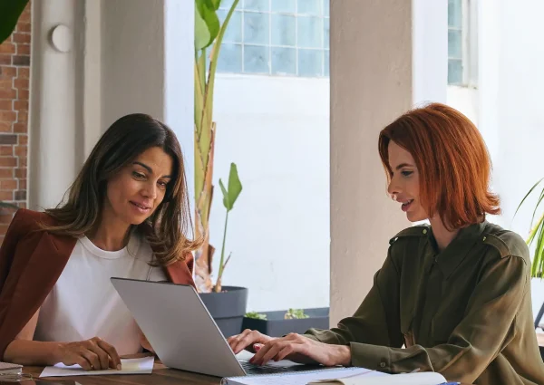 Two people sitting at a table working together on a laptop, reviewing notes and business materials in a bright workspace with indoor plants.